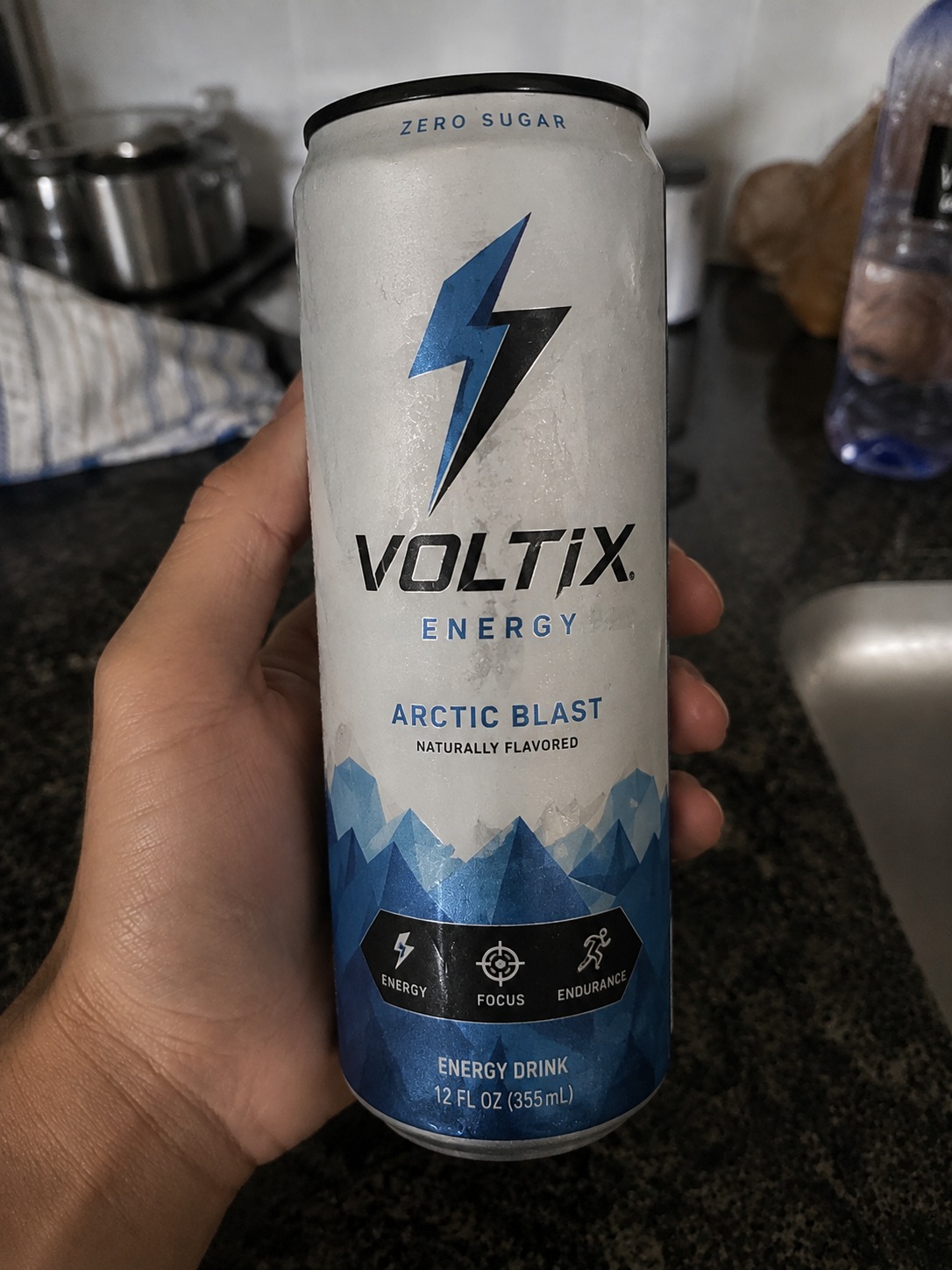 Phone photo of a hand holding a Voltix Arctic Blast energy drink can on a kitchen counter